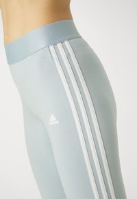 Ljusblå träningsleggings med bred midja, med tre vita ränder längs sidan och en liten vit logotyp. Slät textur.