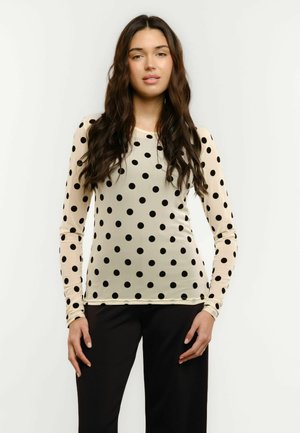 Jeune femme aux cheveux longs et foncés, portant un haut beige à manches longues avec des pois noirs et un pantalon noir, debout devant un fond blanc.