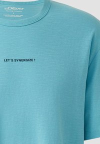 Hellblaues s.Oliver T-Shirt mit schwarzem Text "LET'S SYNERGIZE!" auf der Brust unterhalb des Kragens gedruckt.
