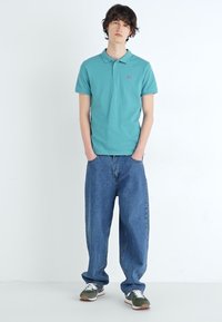 Camisa polo turquesa com colarinho e um pequeno logo, combinada com jeans de ganga azul largos. Footwear com detalhes em verde e branco.