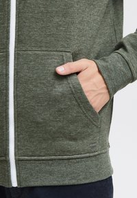 Groene zip-up hoodie van textiel met een witte rits en zijzakken. Heeft ribbels aan de manchetten en onderrand. Kleine logo-detail aan de onderkant.