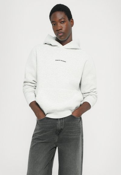 Sweatshirt à capuche gris clair avec poche avant, arborant un texte noir. Associé à un jean gris foncé, affichant un style décontracté.