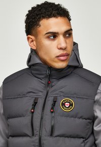 Grijze puffervest met een afneembare capuchon, voorzien van zwarte ritsen, voorzakken en een ronde logopatch op de borst.
