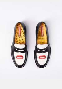 Crno-bijele mokasine s žutim ulošcima s točkicama i dizajnom usana na prednjem dijelu s prikazanim zubima i plavim slovima.