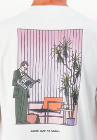 Homem em fato a ler um jornal ao lado de uma cadeira moderna, mesa de apoio e uma planta alta em vaso, com um fundo rosa de riscas verticais.