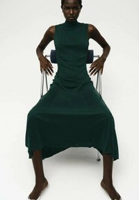 Abito senza maniche verde con scollo alto, texture morbida e silhouette drappeggiata, abbinato a una sedia minimalista e styling a piedi nudi.