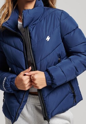 Persoon draagt een blauwe gewatteerde donsjas met ritssluitingen aan de zakken en stelt de ritssluiting aan de voorkant bij, met een wit bliksemschichtlogo op de borst.