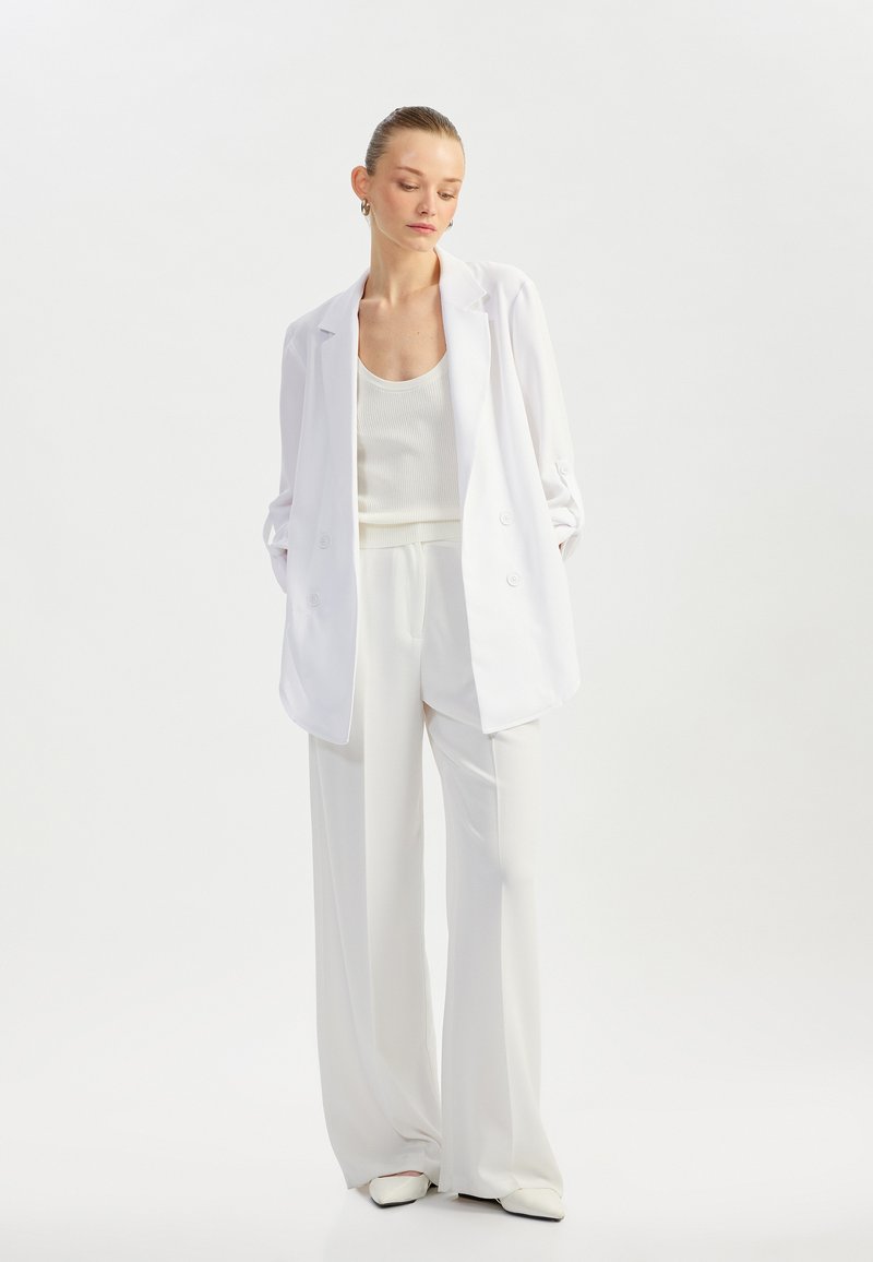 Blazer blanco de doble botonadura sobre un top de tirantes acanalado blanco y pantalones de pierna ancha. Textura suave, diseño minimalista y esquema de colores a juego.