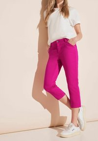 Różowe spodnie o kroju krótszym, z przednimi kieszeniami i zamkiem błyskawicznym z przodu, w połączeniu z białą, fakturowaną bluzką i białymi trampkami. Gładkie wykończenie materiału.