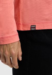 Male hand beside the hem of a pink long-sleeve shirt with a black "Camp Green David" label, paired with black pants.