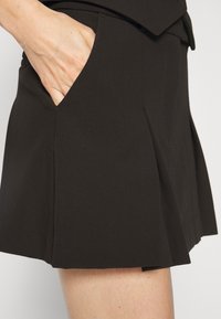 Pantaloni corti neri plissettati realizzati in tessuto morbido, dotati di tasche laterali funzionali e un design pulito e sartoriale. Tessuto liscio senza fantasie.