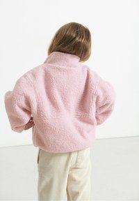 Chaqueta de forro polar rosa con cuello alto y superficie texturizada, llevada sobre pantalones de pana crema. La vista posterior muestra un ajuste holgado y sobredimensionado.
