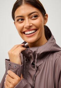 Chaqueta ligera, impermeable, de color malva, con un alto cuello, cremallera frontal y cordón ajustable. Presenta puños de botones y una textura suave.