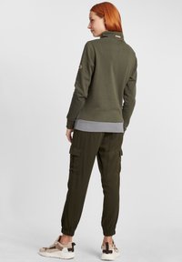 Pullover verde oliva con colletto alto, fondo grigio; abbinato a pantaloni cargo oliva e sneakers beige con sovrapposizione a fantasia.