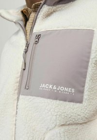 Bijela polar jakna s sivim panelom. Sadrži džep na patentni zatvarač na prsima i otisak "JACK & JONES" u bijeloj boji. Teksturna tkanina, opušteni kroj.