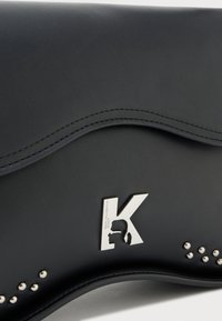 Clutch de couro preto com o logótipo prateado "K", apresentando uma silhueta e pequenos tachas prateadas ao longo das bordas curvas.