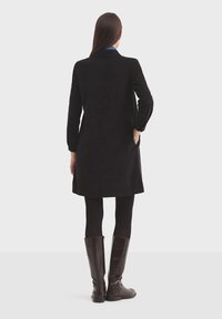 Čierny zamikový kabát s dlhými rukávmi a rovnou siluetou, s bočnými vreckami. Nosený cez čierne legíny a hnedé čižmy po kolená.
