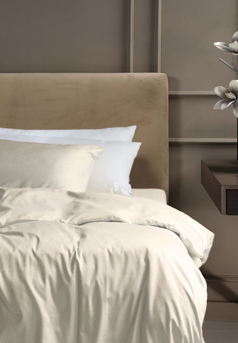 Bettwäsche-Set mit einer cremefarbenen Bettdecke und Kissen, das eine glatte Textur aufweist. Im Hintergrund ist ein beigefarbener Kopfteil und ein dunkler Nachttisch zu sehen.