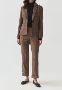 Ruskea tweedmidjekki, istuva malli, yksirivinen nappikiinnitys. Yhdistetty samanvärisiin ruskeisiin housuihin. Musta pooloneule ja mustat ballerinakengät täydentävät asun.