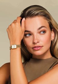 Gouden polshorloge met een vierkante wijzerplaat en een schakelschakelband. Het horloge heeft een witte wijzerplaat en wordt gedragen op een gebruinde pols, met een neutrale top.