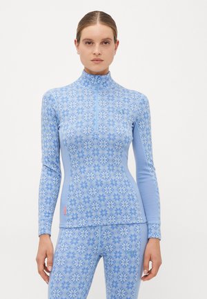 Femme portant un haut à manches longues à motifs bleu clair assorti avec une demi-fermeture éclair et un legging, se tenant devant un fond blanc uni.
