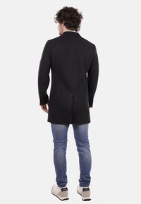 Abrigo negro a medida con un diseño estructurado, con una abertura sencilla en la parte de atrás, combinado con jeans de mezclilla azules y zapatillas de color claro.