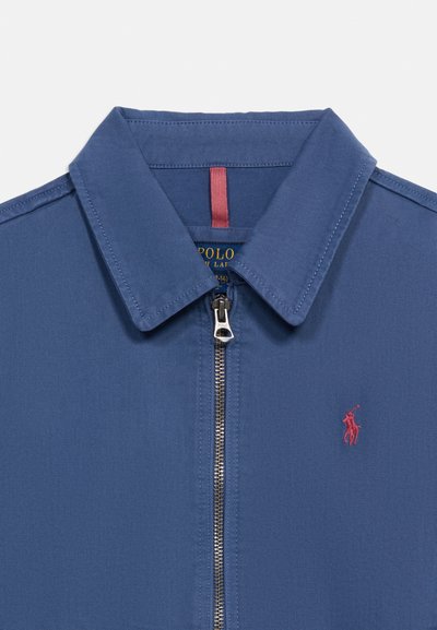 Μπλε μπουφάν με φερμουάρ, μυτερό γιακά, ασημένιο τράβηγμα φερμουάρ και μικρό κόκκινο κεντητό λογότυπο παίκτη πόλο στο στήθος.