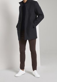 Cappotto di lana navy con colletto alto, chiusura con bottoni e tasche laterali, abbinato a pantaloni marroni scuri e sneakers bianche.