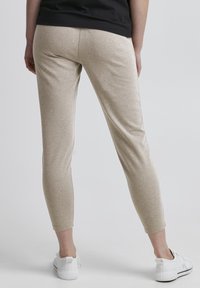 Leggings beiges, extensibles et ajustés, présentant une texture lisse. Associés à des chaussures de sport blanches, sur un fond gris clair.