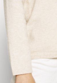Maglione in cashmere color crema con una texture morbida, caratterizzato da orli e polsini a costine. Un primo piano della manica illustra la forma del capo.