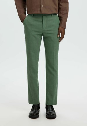 Άνδρας με ραμμένο πράσινο παντελόνι, καφέ πουκαμίσα με κουμπιά, μαύρα δερμάτινα loafers, στέκεται με τα χέρια στις τσέπες σε λευκό φόντο.