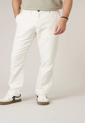 Homme portant un pantalon écru et des baskets blanches à rayures noires, debout les mains dans les poches devant un fond uni.