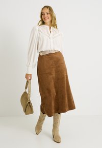 Blusa blanca de manga larga con botones al frente y detalle de encaje, combinada con una falda midi de ante marrón. La modelo lleva un bolso verde y botas vaqueras beige.