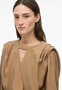 Blouse marron en soie avec un décolleté en V, des détails plissés aux épaules et une texture douce et lisse. Le design est simple et élégant.