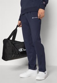 Marinefarbene Jogginghosen mit elastischem Bund, besticktem Logo, kombiniert mit einer schwarzen Sporttasche und weißen Sneakers.