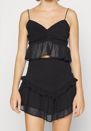 Kobieta ubrana w czarny krótki top na cienkich ramiączkach z marszczonym górnym partem i falbanką u dołu, połączony z pasującą, marszczoną mini spódniczką.