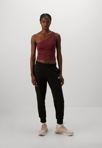 Vrouw staat tegen een neutrale achtergrond en draagt een bordeaux kleurige een-schouder crop top, zwarte joggers, witte sokken en roze sportieve sneakers.