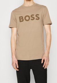 Beige bomullst-shirt med stor brun "BOSS"-texttryck på framsidan. Rund halsringning och korta ärmar, bärs med svarta texturerade byxor.
