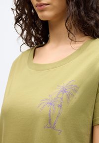 Γυναίκα που φοράει πουκάμισο ελιάς με μοτίβο από μοβ φοινικόδεντρα και καλλιγραφικό κείμενο στο στήθος, με σγουρά μαλλιά που φαίνονται στους ώμους.