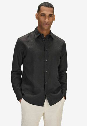 Mand står med hænderne i lommerne, iført en sort skjorte med knapper og lyse bukser mod en ensfarvet hvid baggrund.