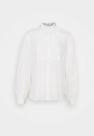 Beige trenssitakki ruutukuvioinnilla, valkoinen ribbineulospaita, vaaleansininen farkkuhame vyöllä ja vaaleanruskeat mokkanahkaiset cowboystövlet.