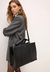 Fekete, strukturált kézitáska texturált bőrrel, dupla fogantyúkkal és levehető vállpánttal. Ezüst színű hardver és logó részletek láthatók.
