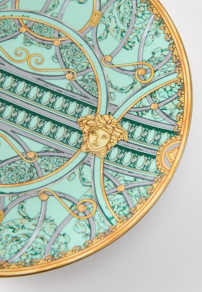 Keramikas šķīvis ar sarežģītu tirkīza un zelta rotājumu, kurā attēloti ziedu motīvi un centrā dekoratīvs medaļons ar sejas dizainu.
