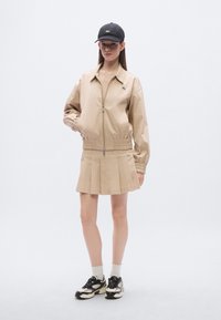 Μπεζ μπουφάν bomber με ελαστικά περιλαίμια, μπεζ φούστα με πιέτες, λευκή κάλτσα με κυματιστή άκρη και μαύρα αθλητικά παπούτσια με κρεμ λεπτομέρειες. Μαύρο καπέλο.