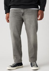 Jeans de mezclilla gris con un corte recto, que presentan una sutil textura desgastada. Combinados con zapatillas de lona negras.