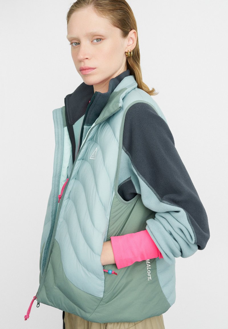 Viegls, daudzslāņu veste teal un zaļās krāsās. Iekļauj quilted dizainu, kontrastējošas pelēkas piedurknes un rozā akcentus ar auklām.