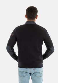 Suéter negro de canalé con parches oscuros en los hombros que contrastan, cuello alto y un diseño ajustado. Se lleva con jeans de color azul claro.