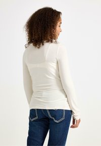 Maglietta aderente a maniche lunghe di colore crema, realizzata in tessuto morbido, abbinata a jeans blu scuro con cuciture in contrasto e un design della tasca posteriore.