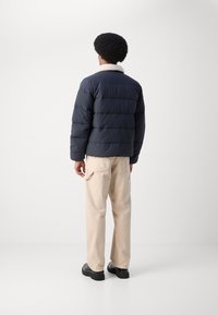 Mørkeblå pufferjakke med horisontale kanaler og en cremet shearlingkrave, sammen med beige bukser og sorte sko, vist fra ryggen.