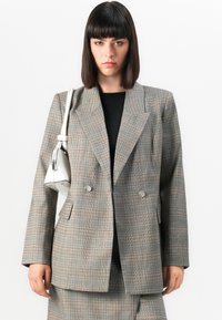 Grå rutete dobbelspent blazer med spisse lapeller, som har to frontlommer og metallknapper, kombinert med en svart topp og en hvit veske.